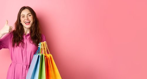 Young happy female shopper showing thumbs up, pleased with good discounts, buying staff on sale, holding shopping bags and smiling pleased, pink background.